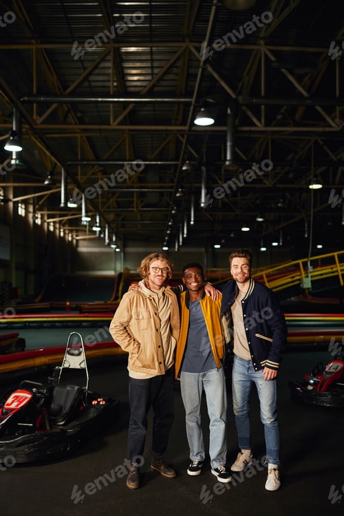 Preview: multicultural male friends smiling and standing near racing cars inside of karting track, smile