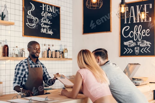 Preview: handsome smiling african american barista giving menu to clients on bar counter in cafe