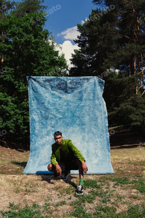 Preview: fashionable african american man sitting on haunches in forest near blue drapery