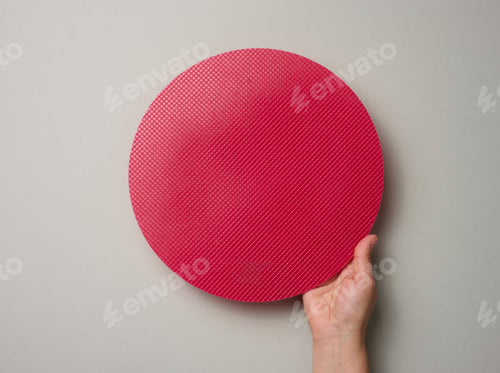 Preview: Female hand holds empty red plastic textured cake plate on a gray background, top view
