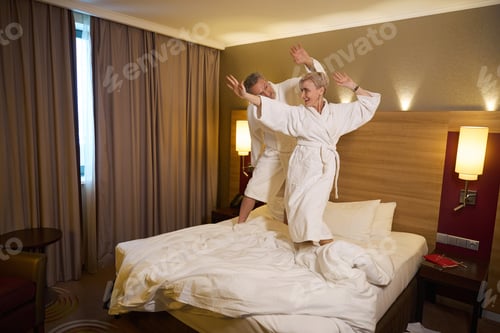 Preview: Joyful adult caucasian couple dancing and celebrating on bed in hotel room