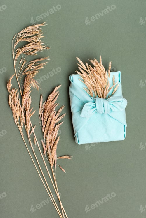 Preview: Eco friendly furoshiki gift with ears of dry grass on green background. Vertical view