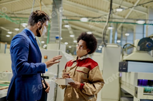 Preview: Black female worker and company manager wearing face masks while talking at woodworking factory.