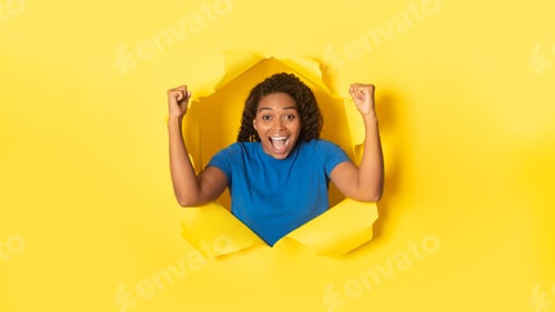 Preview: Overjoyed black woman cheering with clenched fists, celebrating victory, posing in torn hole of