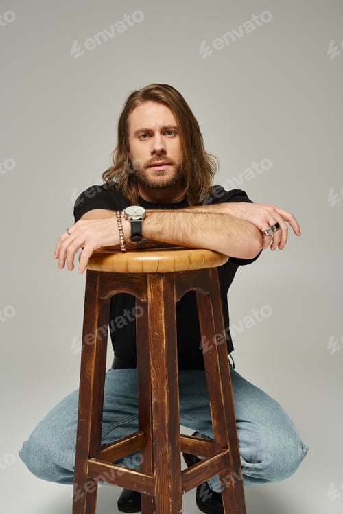 Preview: bearded and handsome man with long hair in jeans and t-shirt posing next to wooden chair on grey