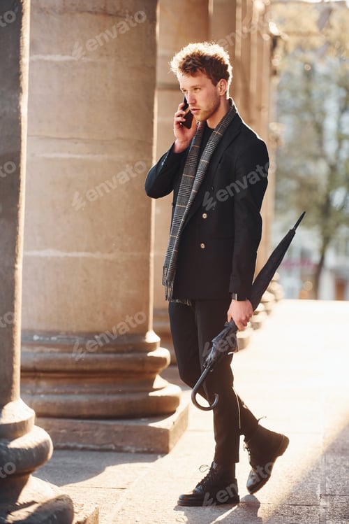 Preview: Elegant young man in formal classy clothes standing and talking by the phone outdoors in the city