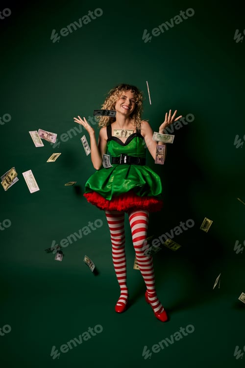 Preview: cheerful curly woman dressed as new year elf standing under money rain cheerfully looking at camera