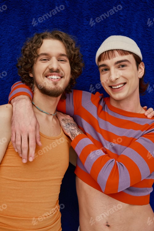 Preview: two contented good looking gay men in vibrant clothes posing on dark blue backdrop, pride month