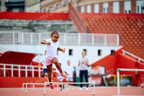 Preview: Black little girl jumping over hurdles on running track at the stadium.