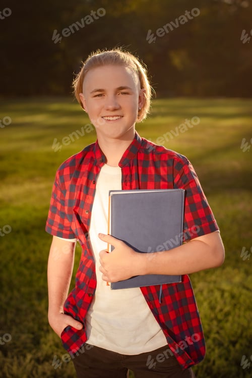 Preview: happy smiling teenager boy with notebook in hands stand on school yard or park in sunny day