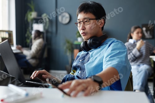Preview: Creative Asian Man with Headphones Using Laptop at Workplace