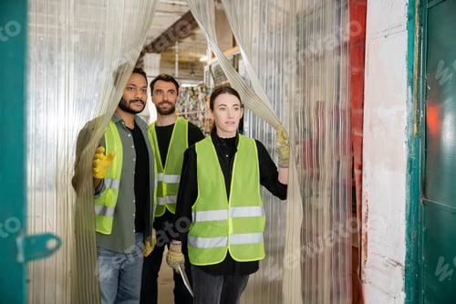 Preview: Young worker in protective gloves and vest holding digital tablet while standing near interracial