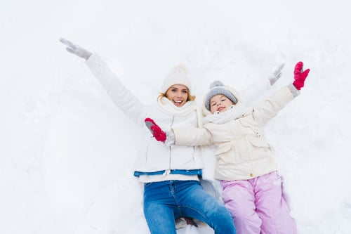 Vorschau: glückliche Mutter und Tochter mit offenen Armen lächeln in die Kamera, während sie zusammen im Schnee liegen