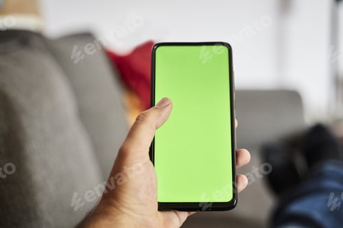 Preview: Closeup shot of a man holding a smartphone with a green screen whilst sitting on a gray sofa