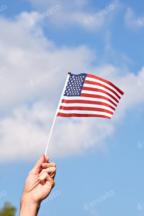 Preview: Raised hand with waving American flag against blue sky