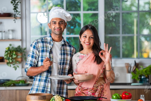 Preview: Indian asian couple promoting, showing or holding kitchen utensils in kitchen