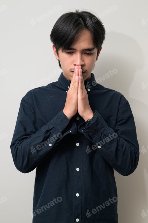 Preview: Portrait of sad and stressed young Asian Thai man praying against white background