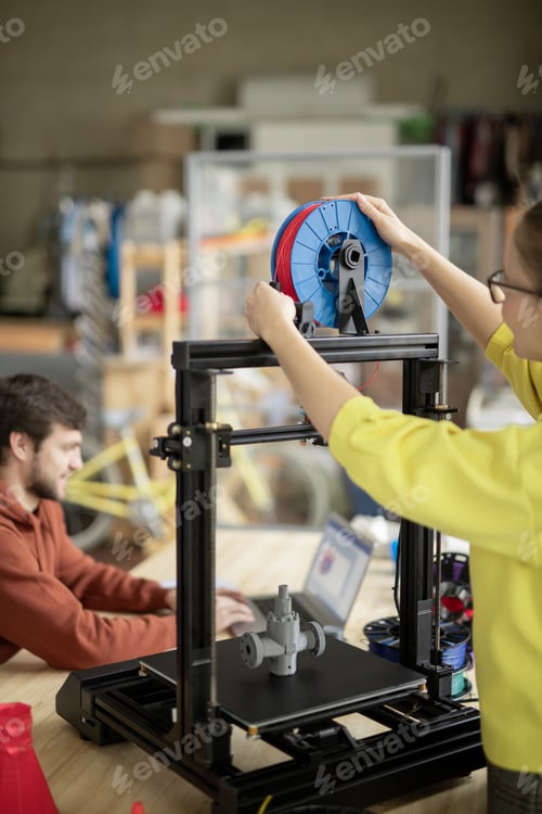 Preview: Young woman putting new spool with filament on 3d printer before starting work