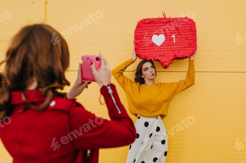 Preview: Woman in yellow sweater posing with heart icon. Outdoor shot of two friends making photos