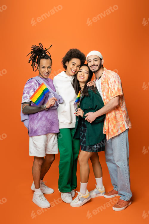 Preview: full length of stylish and cheerful multicultural friends embracing while holding lgbt flags on