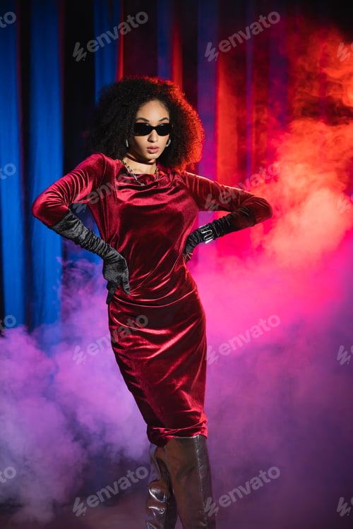 Preview: Elegant african american woman in velvet dress and gloves posing on red background with blue light