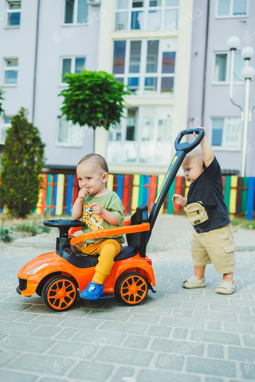 Preview: A one-year-old boy pushes a big toy car while playing outside. Children are playing in the street