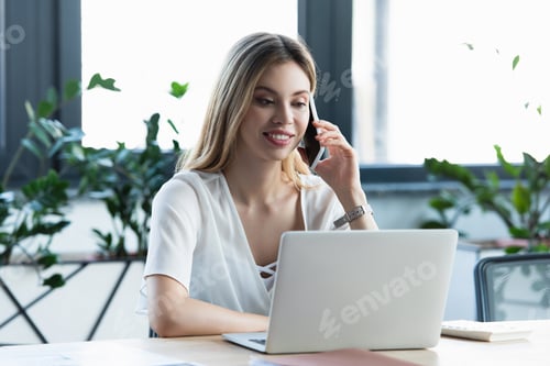 Preview: Cheerful businesswoman talking on mobile phone and using laptop in office