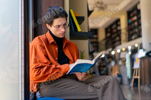 Preview: a young guy with glasses is sitting in library cafe and carefully reading a book about hobbies