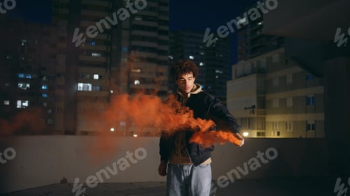 Preview: Hipster holding smoke bomb at night city. Curly guy teenager burning red grenade
