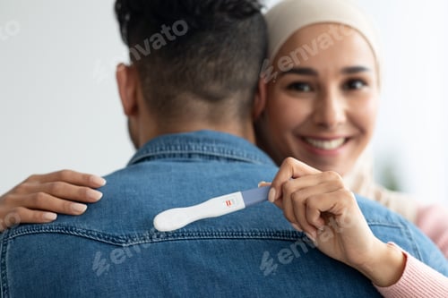 Preview: Cheerful muslim woman with pregnancy test hugging husband, closeup
