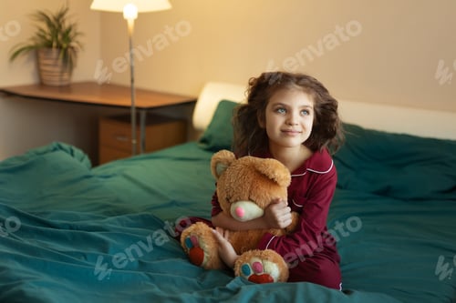 Preview: 7 year old girl in red pyjamas sits in green bed with her teddy bear toy at home