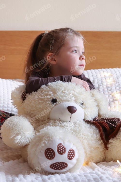 Preview: A little pretty girl looks with a bored look. A teddy bear sits nearby. Bored mood.