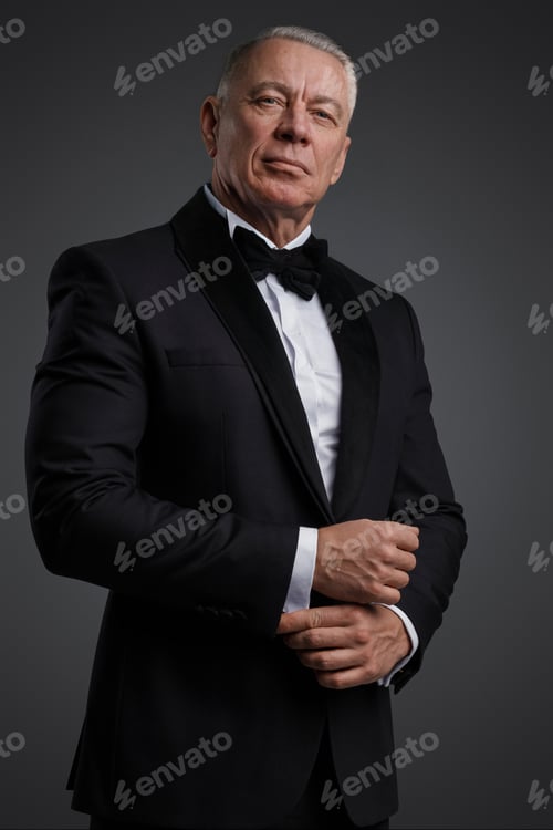 Preview: The distinguished old man stands confidently in a black suit and bow tie against a grey backdrop
