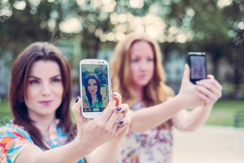 Preview: Two young female friends taking selfie on smartphones in unison