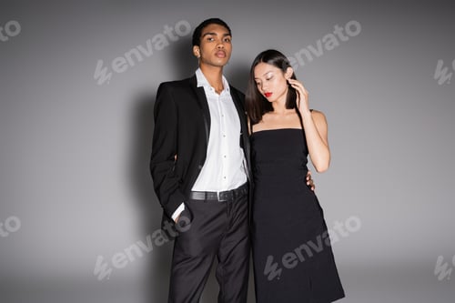 Preview: african american man in elegant suit looking at camera near asian woman in black dress on grey