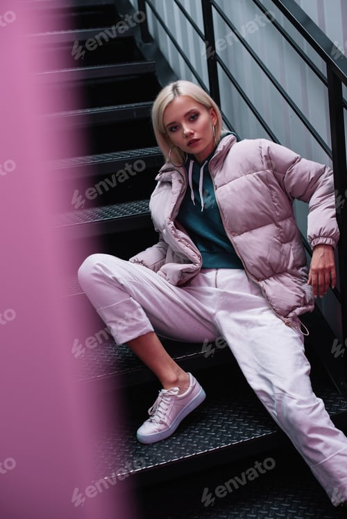 Preview: blonde girl in down jacket sitting on stairs and looking at camera