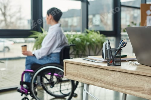 Preview: focus on table with stationery with blurred disabled woman in wheelchair on backdrop in office