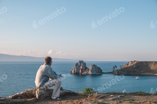 Preview: A middle-aged man is sitting on the shore of Lake Baikal and enjoying the summer landscape.