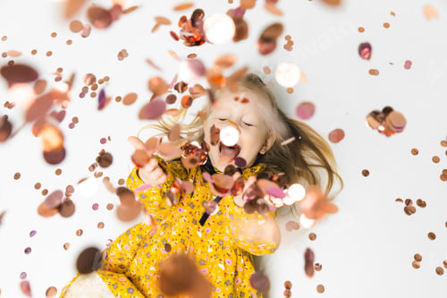 Preview: Happy little blonde girl in yellow dress catching confetti, having fun on white background
