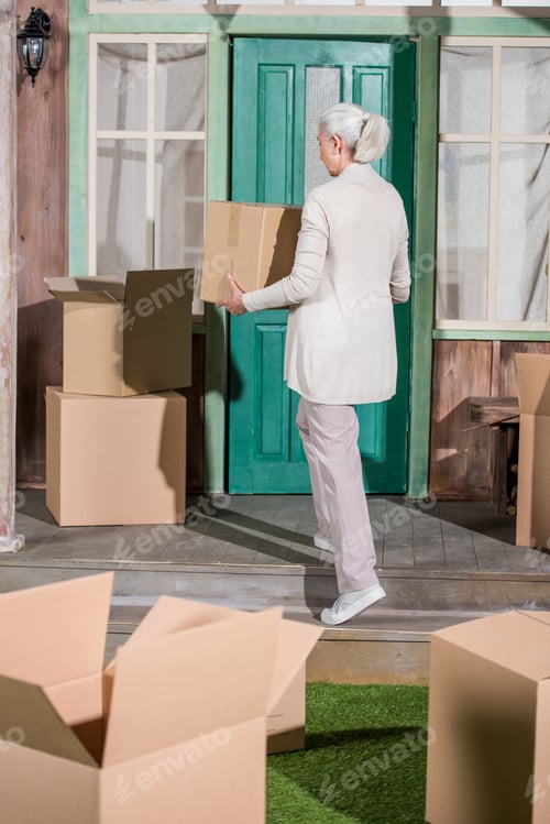 Preview: Back view of senior woman holding box while standing on porch of new house with cardboard boxes