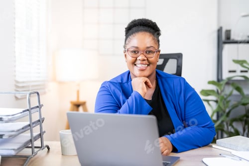 Preview: Smiling african american businesswoman in blue blazer working on laptop at office desk.