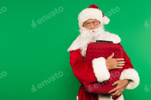 Preview: Santa claus in hat and eyeglasses hugging suitcase isolated on green