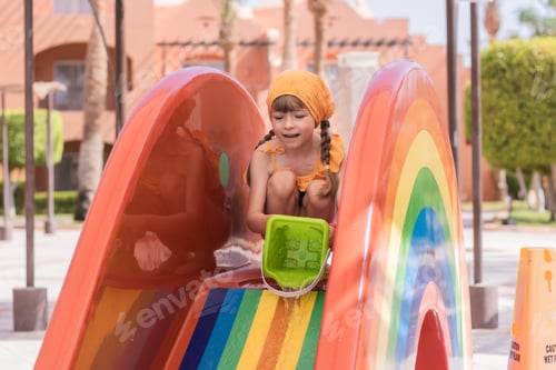 Preview: Little girl in a swimsuit and a scarf from the sun plays with water on a slide near the pool