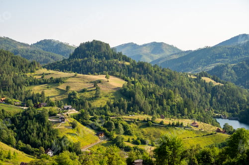 Preview: Lush green valley with rural houses surrounded by forested mountains under clear blue sky.