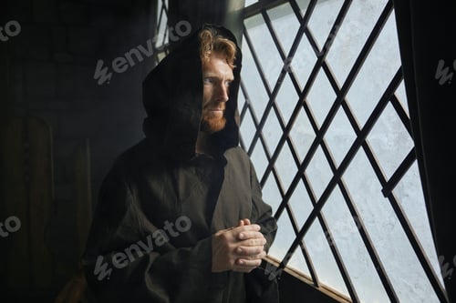 Preview: Pensive monk in medieval costume with hood looking through window of monastery
