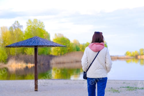 Preview: Slender woman with dark hair walks at the bank of the river. Spring green trees and calm water.