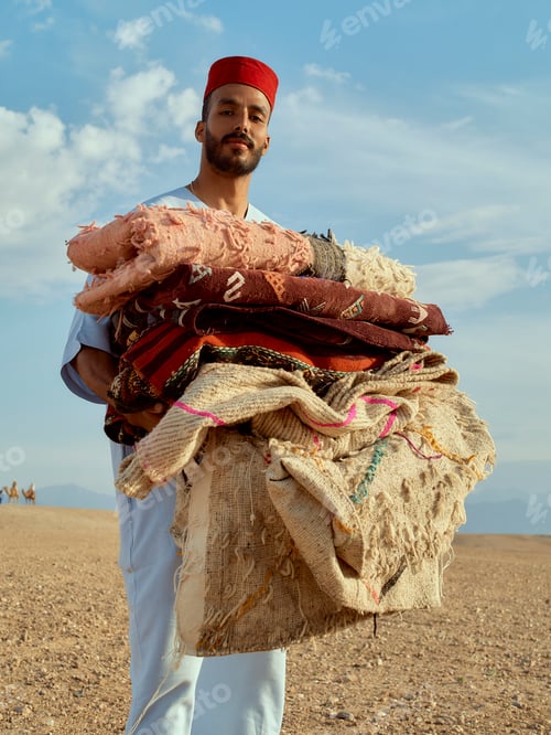 Preview: Man standing in a desert landscape holding a stack of colorful carpets.
