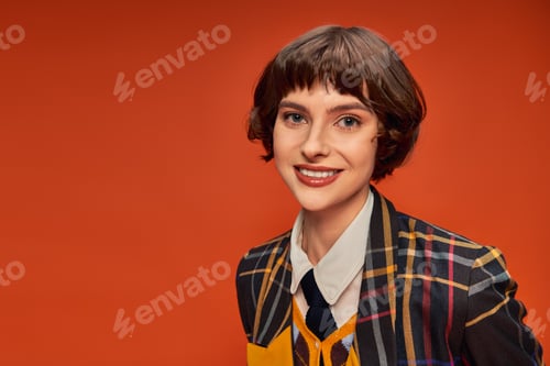 Preview: optimistic and young student girl in checkered college uniform smiling on orange background