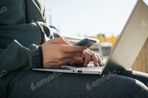 Preview: Close-up of male hands using mobile phone and laptop outdoors. Male freelancer browsing internet