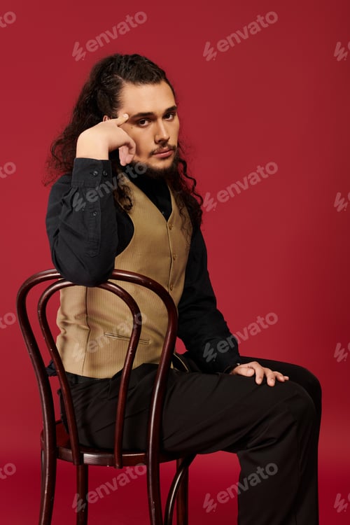 Preview: Captivating young man in stylish vest sitting thoughtfully on a chair against a vibrant red backdrop